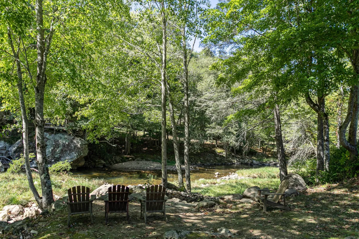 This image captures a pair of wooden chairs placed beside a serene, gently flowing river, bordered by lush green trees. The tranquil scene invites relaxation and offers a strong sense of connection to the natural surroundings, invoking the peacefulness of riverside pursuits such as fishing or simply unwinding while immersed in nature.