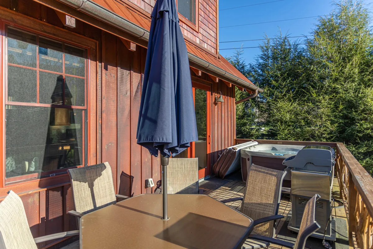 A spacious deck area featuring an inviting outdoor dining table with a parasol, a modern hot tub, a gas grill, and lush greenery surrounding the space. The layout fosters both relaxation and social gatherings, with the hot tub and grill easily accessible from the dining area.