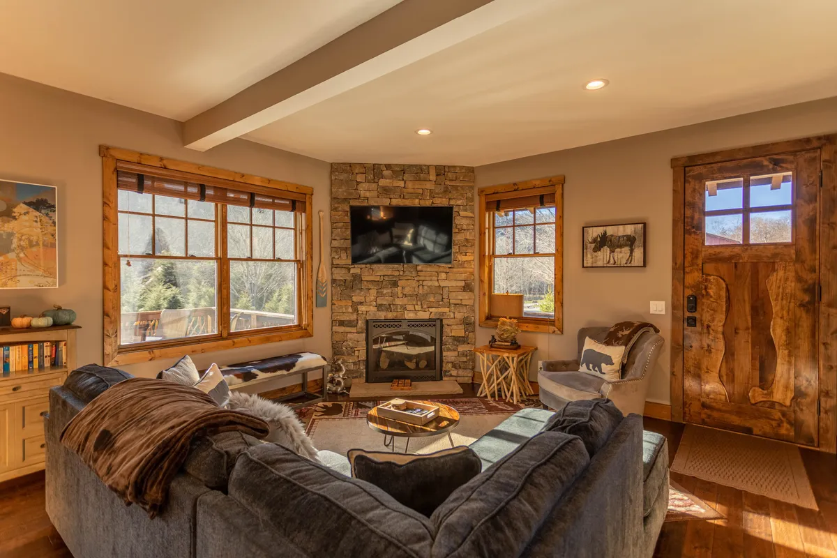 A spacious, inviting living room centered around a striking stone fireplace, featuring a large sectional sofa, wooden coffee table, and expansive windows that flood the space with natural light. Custom artwork and mountain-inspired décor adorn the walls, enhancing the cozy yet sophisticated feel. The arrangement encourages relaxation, conversation, and family gatherings.
