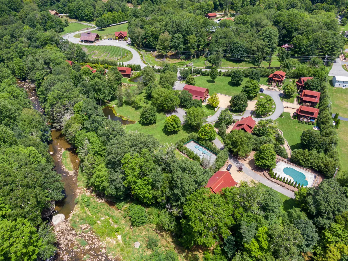 A sweeping exterior view showing a winding river, red-roofed cabins, lush greenery, a swimming pool, and landscaped gardens. The scene illustrates a harmonious blend of nature and resort living, with families swimming, relaxing, and picnicking by the water.