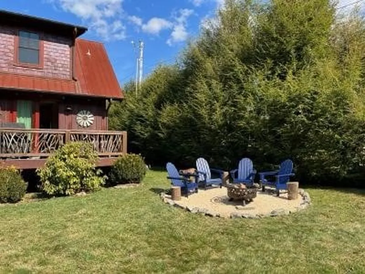A cozy fire pit area with blue Adirondack chairs arranged around a stone-surrounded fire pit, set in front of a rustic cabin with a red metal roof and lush greenery. The inviting setup encourages evening gatherings, storytelling, and stargazing.