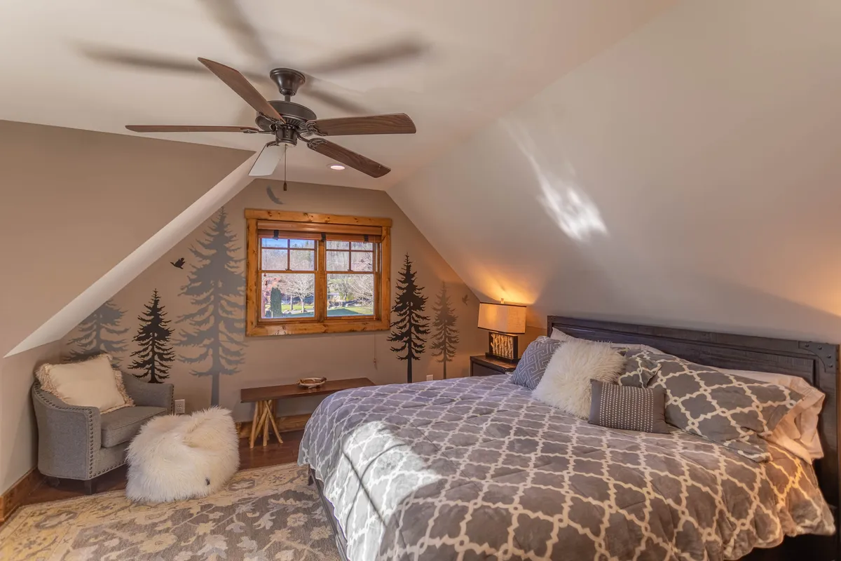 A serene bedroom with a bed featuring patterned linens, a wooden-framed window letting in ample natural light, a wall mural of trees, and a comfortable armchair perfect for reading or relaxing.