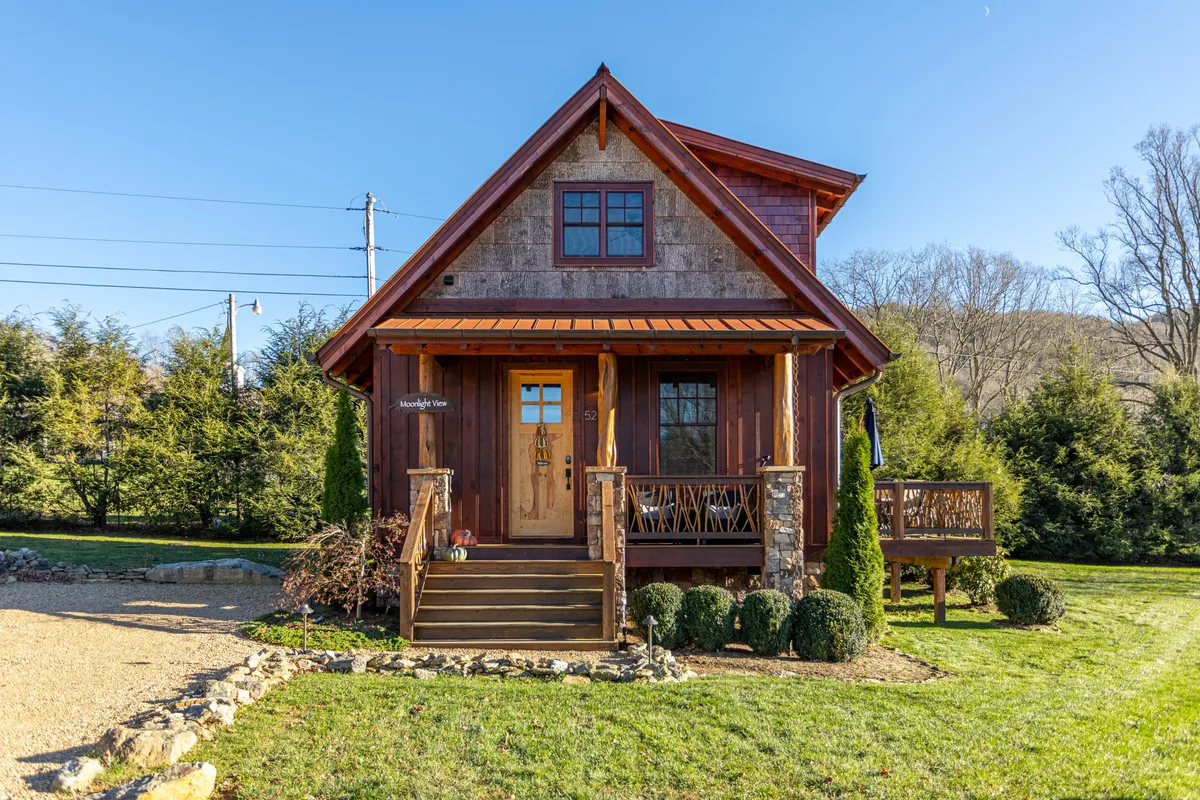 The image showcases the inviting front porch of a luxury mountain cabin, complete with a sturdy wooden door, charming stone steps, and a beautifully landscaped garden. The scene is warmly lit and surrounded by natural beauty, setting a cozy, welcoming atmosphere perfect for relaxing and enjoying the tranquil mountain environment.