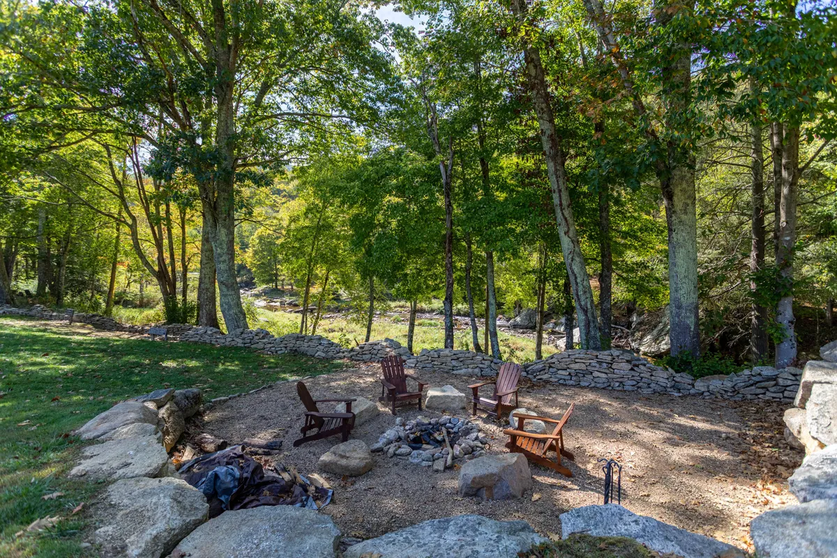 An outdoor fire pit surrounded by Adirondack chairs and a low stone wall, encircled by trees. The space is perfect for cozy, intimate gatherings, stargazing, and sharing stories by the fire under open skies.