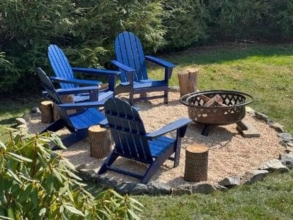 A close-up of blue Adirondack chairs arranged around a glowing fire pit surrounded by greenery. The setup invites families to gather, share stories, and enjoy classic campfire moments under the stars.